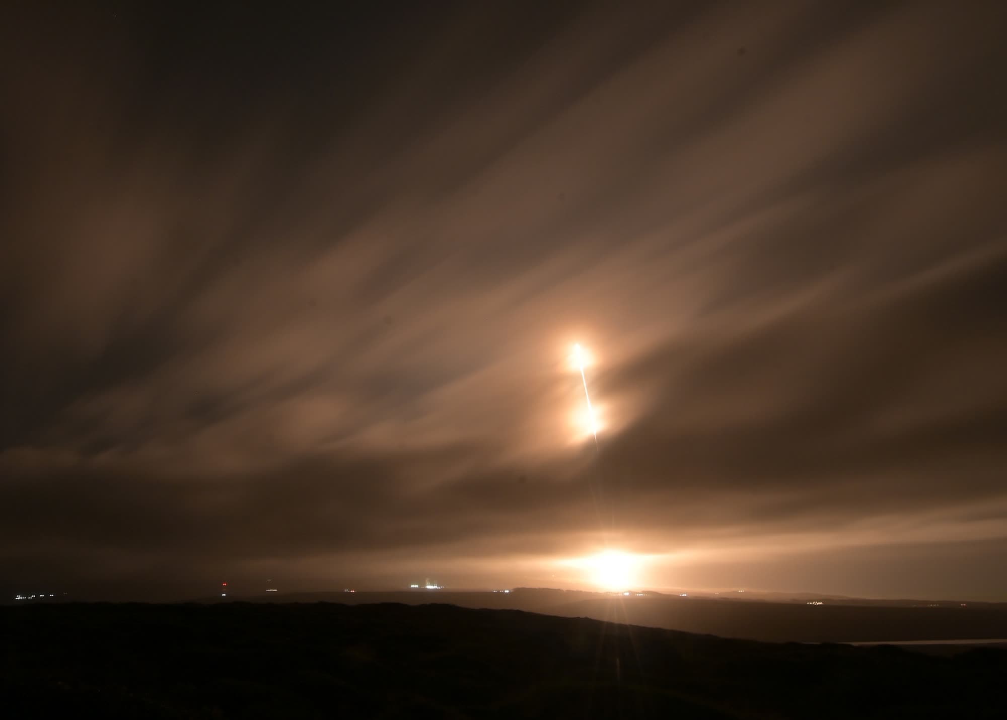 Long-exposure photo of a Falcon 9 launch arcing over the dark Vandenberg coastline, its trail glowing orange